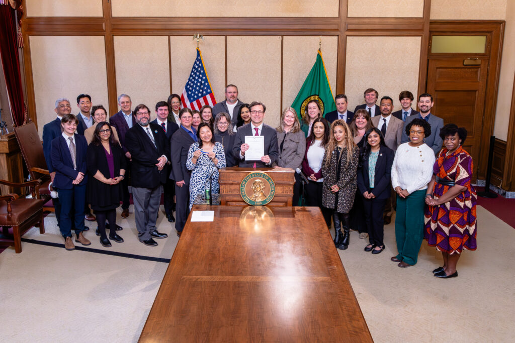 A diverse group of people stand together behind a podium with the Washington state seal on it