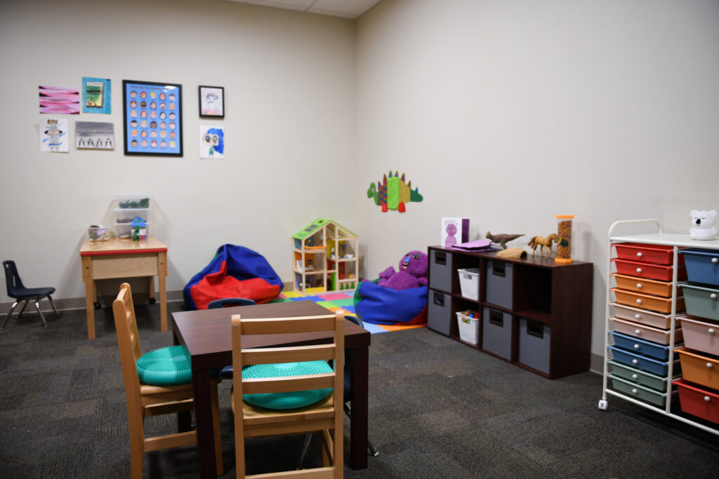 A colorful play therapy room for children with a table and chairs a doll house that sits in the corner and a purple plushie rests on a bean bag chair