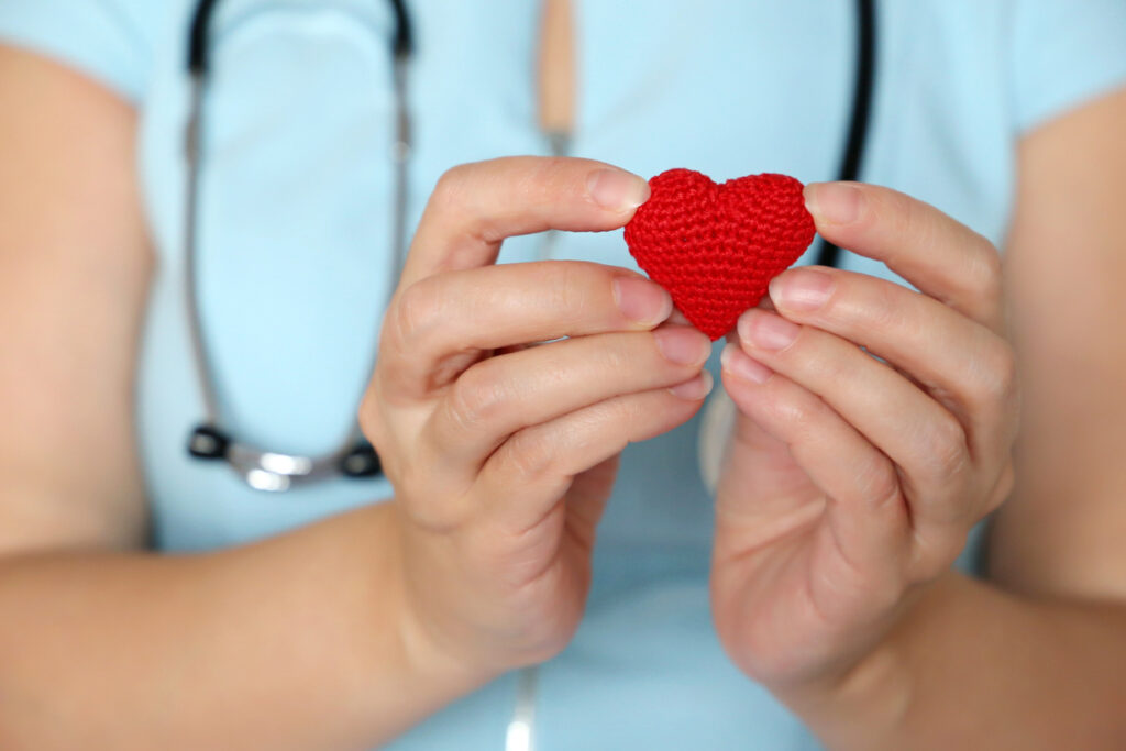 A health care worker in blue scrubs, with a stethoscope draped around her neck, holds a small red crocheted heart.