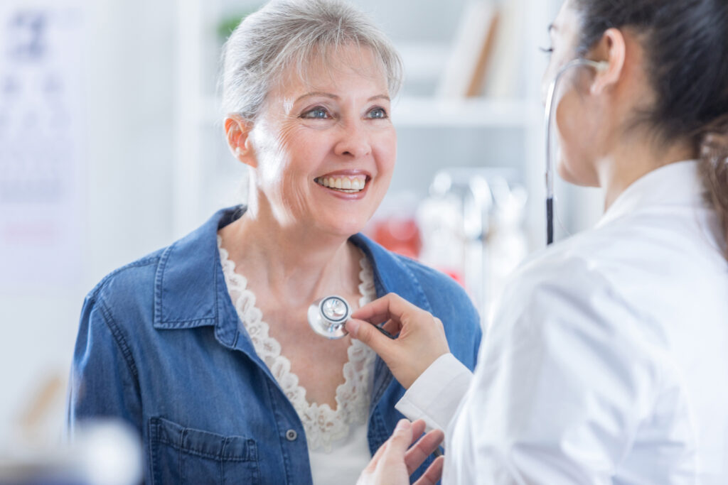 Physician listens to senior Caucasian woman's heartbeat with a stethoscope. The woman is smiling at the doctor.