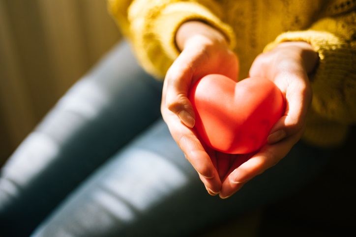 A person ina yellow sweater and blue jeans holds a small red heart in their hands. 