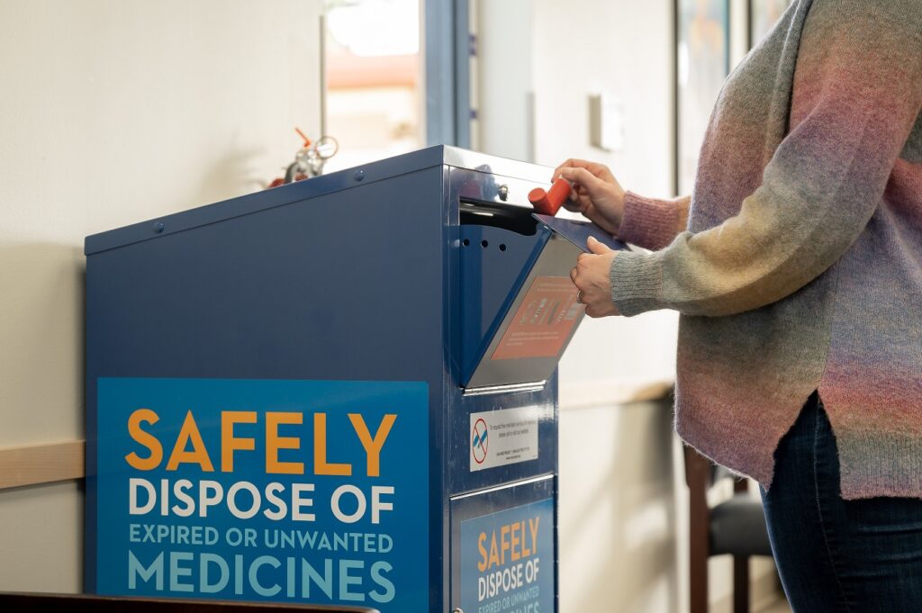 A person in a pastel sweater drops a prescription bottle into a large secure metal blue bin witha sign reading "Safely dispose of unwanted or unused medicines."