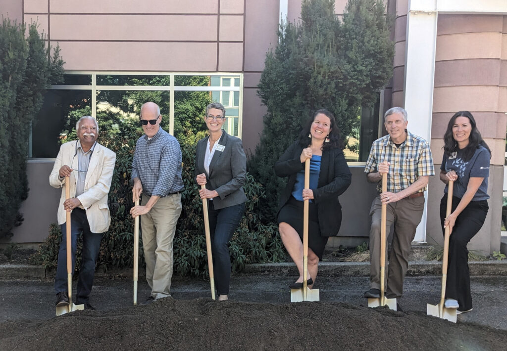 A group of six people stand in front of a building with their feet positioned on top of shovels in a a pile of soil 