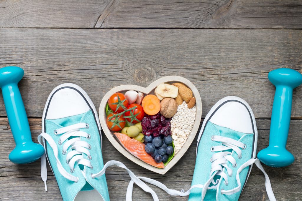 A heart shaped bowl full of healthy food with a pair of turquoise tennis shoes and a set of turquoise weights set beside it. 