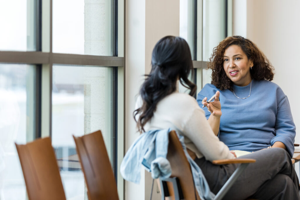 A healthcare worker sits with a patient in front of a bank of windows and discusses her care